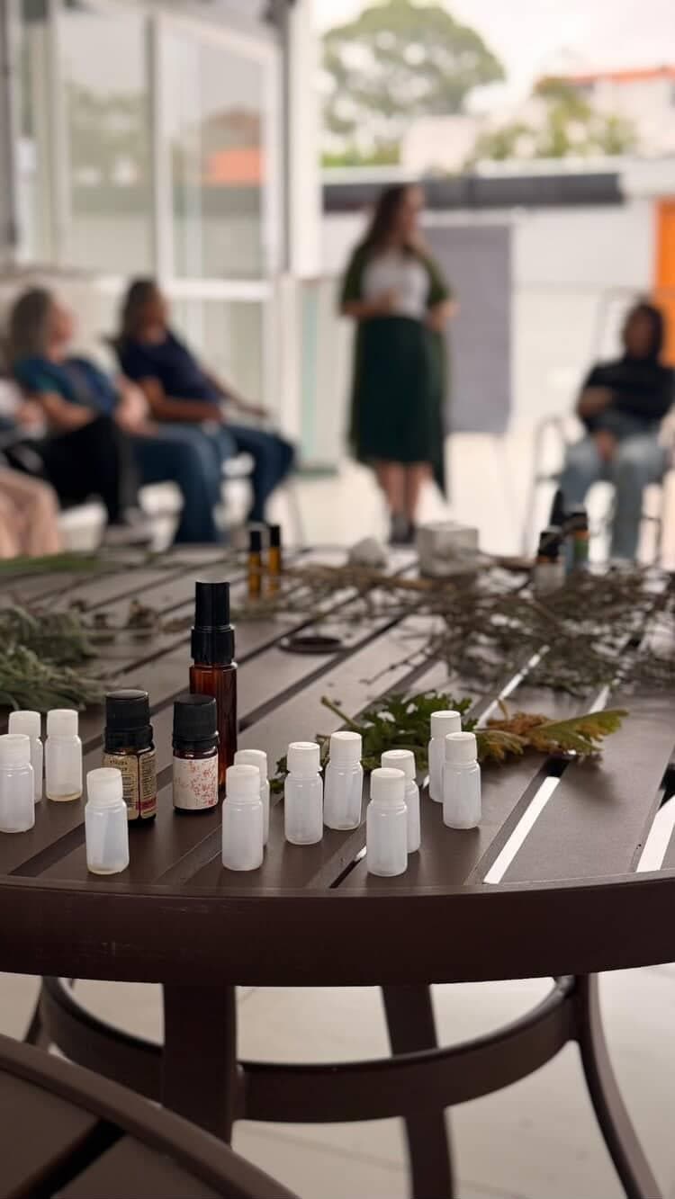 Frascos de óleos essenciais e ramos de ervas sobre a mesa, com participantes sentados ao fundo